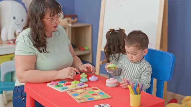 Woman guiding little boy stacking colorful wooden rings with hands at a red table inside a building play area; early learning curiosity.