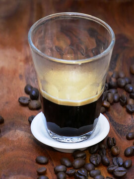 A clear glass of dark coffee with crema sits on a saucer surrounded by coffee beans.