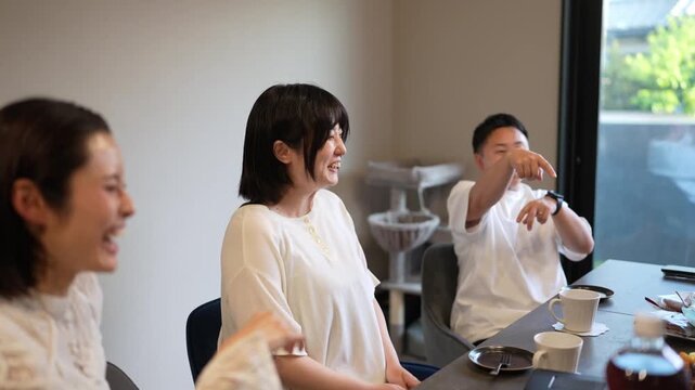 Three people in a bright, modern room. They sit around a table with drinks and cups. One person speaks, smiling, as others listen. They share a relaxed, pleasant conversation