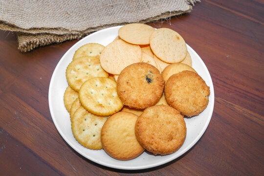 Assorted cookies and biscuits on a white plate on a wooden table. A collection of sweet baked snacks including chocolate cookies, butter biscuits, crackers, and sandwich cookies on a plate.