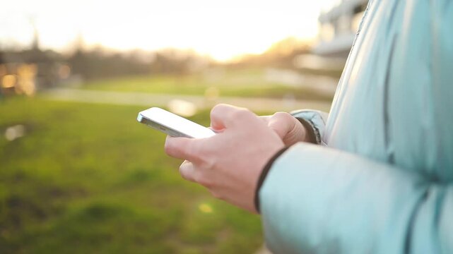 a person is seen texting on a phone while walking on an outdoor pathway surrounded by grass and buildings in the background, creating a neutral mood and showcasing an urban lifestyle.