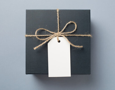 Rectangular dark charcoal gray gift box wrapped with rustic natural jute twine string and a blank white paper tag, presented on a muted monochrome bluish gray background from a top-down view