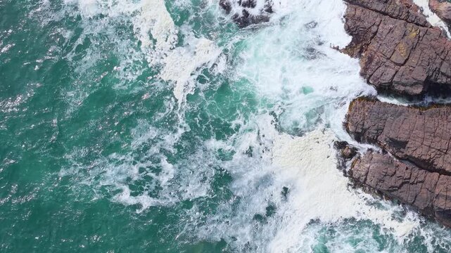 Cinematic top-down static aerial view of powerful turquoise ocean waves and white foam breaking against rugged rocky cliffs on the wild coast of Chilo&eacute; Island in Chilean Patagonia.