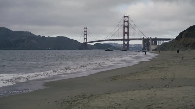 Wide Shot Of Waves Crashing On Shore Near Golden Gate Bridge With Foreground People Walking