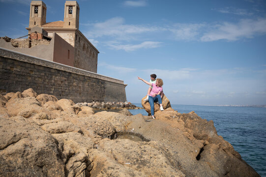 Middle aged woman and her teenage son admiring the historic church from the rocky shoreline while exploring Tabarca Island during a summer vacation in Spain