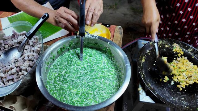Thai street food vendor rapidly stirs scrambled eggs with diced taro and chopped green onions on a large hot flat griddle during the final cooking stage at a local night market. Authentic Asian street
