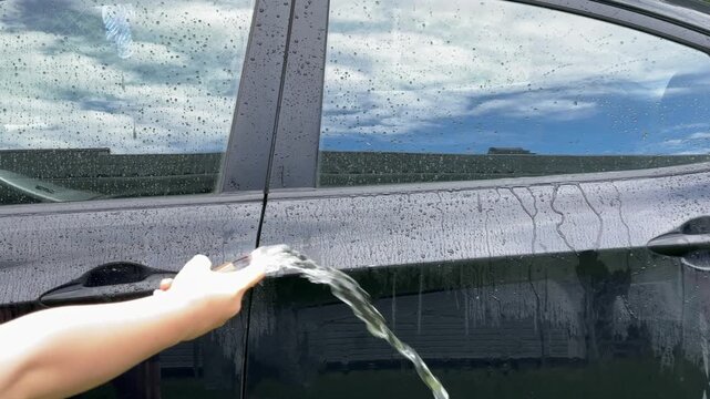  A hand pours water from a clear cup onto a dark car door to test the hydrophobic coating outdoors. car wash, water repellent, auto detailing.