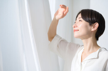 カーテンを開けて朝日を浴びる日本人女性のビューティイメージ