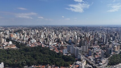 Obraz premium Visão aérea da cidade de Campinas, SP, com prédios altos, bairros densos, áreas verdes e horizonte urbano amplo sob céu azul, mostrando a expansão da metrópole no interior paulista.