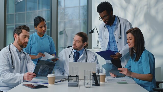 Medical team cooperating working together in clinic. Caucasian doctor boss leader confident talk leading medicine conference with multiethnic five diverse people doctors nurses at meeting in hospital