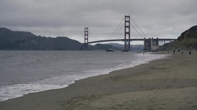 Golden Gate Bridge, Sandy Beach Foreground With People Walking - Wide Shot And Overcast Sky