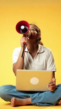 Girl Using Laptop with Megaphone While Sitting Against Yellow Seamless Background Making Online Announcement or Promotion, Smart Indian Asian Child Promoting Educational or Digital Content Loudly