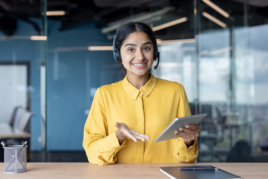 Portrait of an Indian woman in a headset sitting in the office in front of the camera and smilingly communicating, holding a tablet in her hands