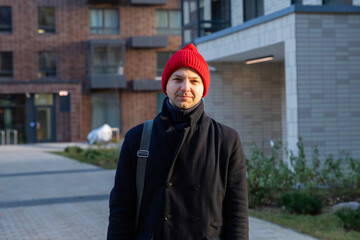 Fototapeta premium Student standing near university dormitory. Young man wearing red beanie and warm coat outdoors. Campus life concept. Education and housing solution for international student in city.