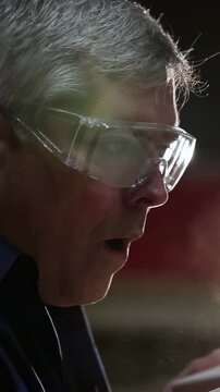 Vertical video: Inspecting senior in dark shirt blowing dust from part at shop bench, with goggles
