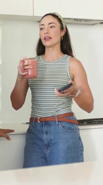 Vertical video: Talking woman in striped top showing phone to hand with pink mug at kitchen counter