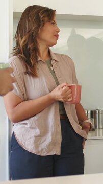 Vertical video: Hearing left speaker, woman in beige shirt leaning at kitchen counter with pink mug