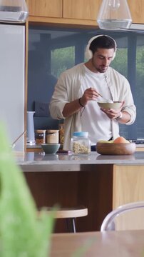 Vertical video: Eating man in white tee listening, using spoon on green bowl at kitchen island