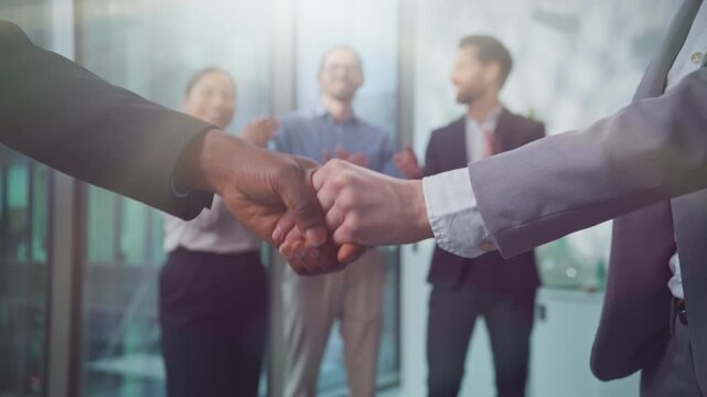 Close up Caucasian and African business men shaking hands near carefree three multiethnic partners applauding joyful in sunny corporate office. Multiracial businessmen handshake trust successful deal