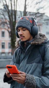 A young adult male wearing a winter jacket with fur lining and a beanie with ears is engrossed in using his cell phone.
