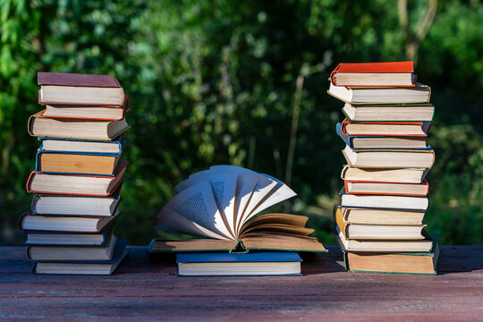 Two stacks of hardcover books flanking an open book with fanned pages on wooden table against blurred green nature background representing education and wisdom