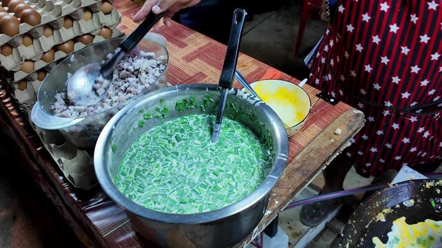 Thai street food vendor pours beaten eggs onto a hot flat griddle over coconut batter with chopped green onions while preparing a traditional street food dish at a local night market in Thailand. Auth