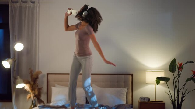 Joyful African American ethnic woman in headphones singing in phone like microphone dance enjoying music moving energetic girl female sing in telephone pretending singer jumping dancing joy on bed