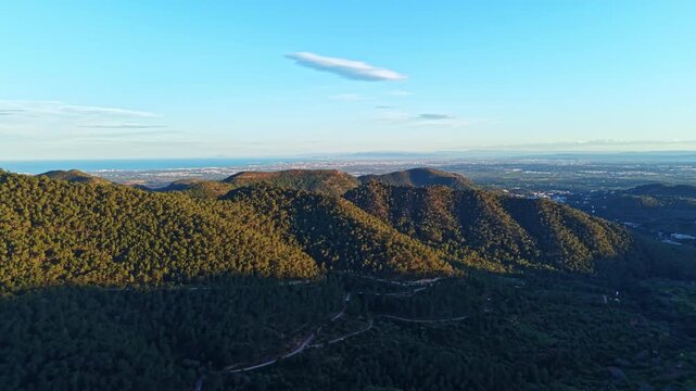 Mountains stretch across the landscape with shadows from the setting sun. The sea glimmers in the distance as the city lights begin to shine.