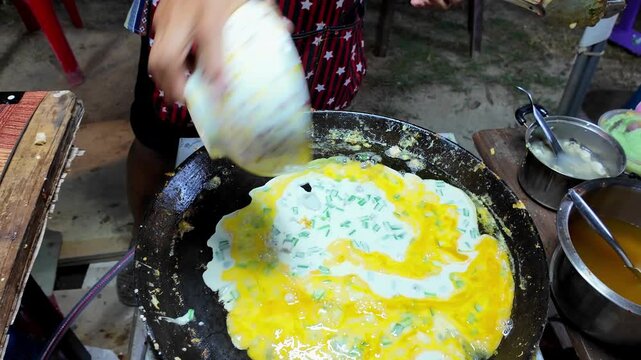 Thai street food vendor finishes cooking scrambled eggs with diced taro and chopped green onions on a large hot flat griddle at a local night market. Authentic Asian street food preparation filmed in 