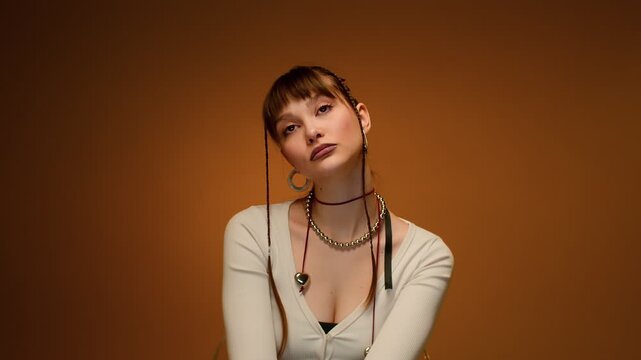 Calm studio portrait of adult woman with warm orange backdrop, bold makeup, braided hair, jewelry