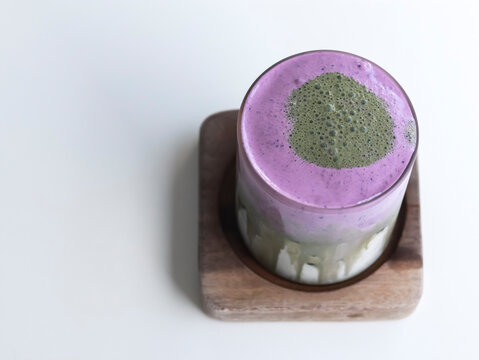 A high-angle, top-down studio shot of a layered matcha and ube smoothie or latte, served in a glass on a square wooden coaster against a clean white background.