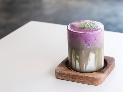 A layered matcha and ube latte, served in a glass on a square wooden coaster against a clean white surface. 