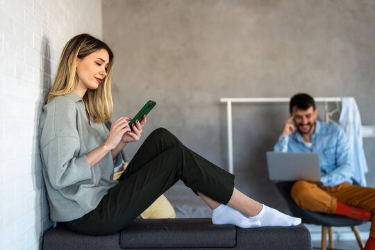 Smiling caucasian woman using her smartphone while her partner works in the background, home office