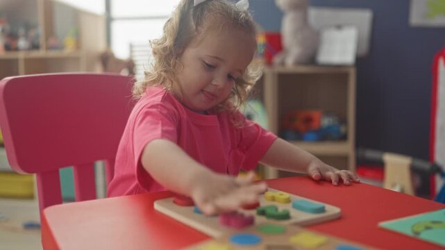 Little girl places a colorful wooden puzzle piece with her right hand on a red table inside a building playroom; concentration.
