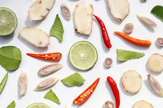 A colorful, close-up pattern of Thai herbs: lime slices, galangal, lemongrass, chilies, and kaffir lime leaves.