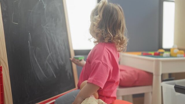 Girl erases chalkboard with green eraser while holding a doll in a playroom; curiosity play learning.