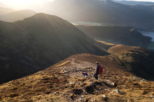 A female hiker and their dog walking down from the summit of Grasmoor with Whiteless Pike and Rannerdale  in the distance on a bright winters morning in the Lake District, UK
