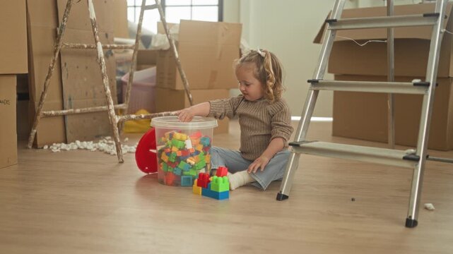 Little girl reaches hand into a clear tub of colorful plastic blocks while sitting among moving boxes and ladders in a building; playful curiosity family relocation.