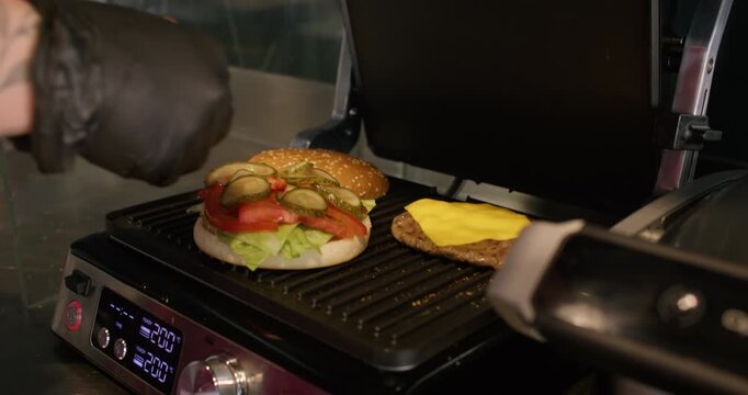 Grilling food. Burger preparation. Gloved chef hands placing pickle slices on bun with lettuce tomato. Roasting beef patty cheese.
