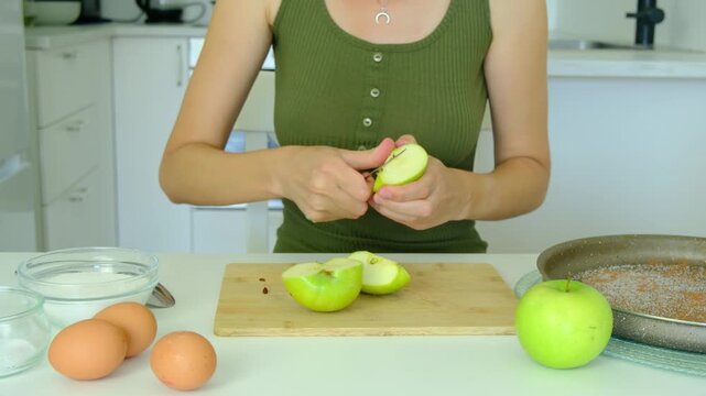 Female hands use knife to slice fresh green apples on wooden board. Sliced fruits go inside round baking pan on white kitchen table next to raw eggs and bowl with white sugar.