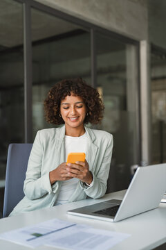 Smiling young indian or muslim arabian businesswoman using smartphone at desk at laptop in office. Mobile work communication. Latin curly young woman working on cellphone phone at workplace. Vertical