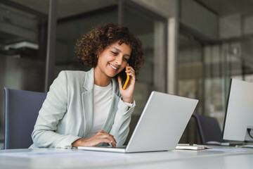 Fototapeta premium Young Indian woman manager ceo using cell phone mobile, laptop for business call. Smiling Latin female businesswoman holding smartphone in office, talking with client, doing professional consultation