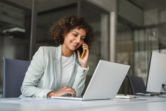 Young Indian woman manager ceo using cell phone mobile, laptop for business call. Smiling Latin female businesswoman holding smartphone in office, talking with client, doing professional consultation