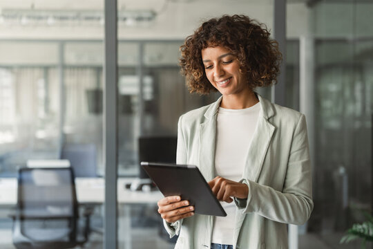 Focused young professional specialist latina hispanic business lady working on tablet pc standing in modern office workplace. Indian IT manager woman using tab computer device app for work online