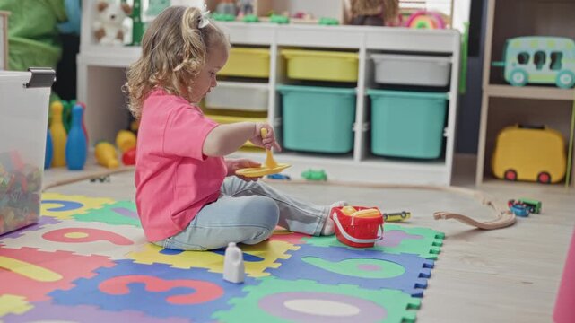 Little girl sitting crosslegged on foam alphabet mat holding a yellow plate and spoon, arm raised, wearing pink shirt and jeans in a building; playful imagination.