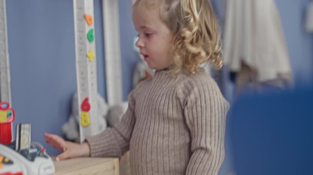 Girl touches a toy cash register and presses buttons on a wooden shelf beside storage bins and a height ruler in a playroom; quiet curiosity.