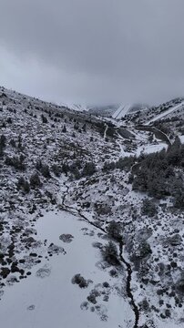Vertical aerial drone footage of Valdesqui ski resort from a distance in the Sierra de Guadarrama, Madrid, Spain, with snowy slopes, a stream in the valley, and misty overcast sky