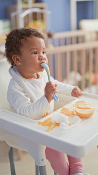 Little girl holds spoon and scoops orange baby food toward mouth at nursery highchair with bowl on messy tray and stained bib; curiosity independence.