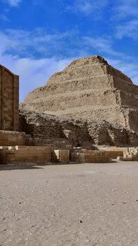 Ground level view of Step Pyramid of Djoser at Saqqara, Egypt, tiered limestone structure with ancient ruined entrance pavilion and stone foundations.