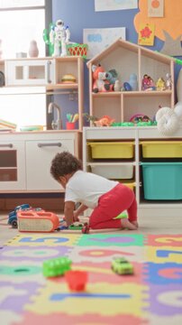 Little girl kneeling, hands reaching for toy cars on foam mat in a playroom building; playful curiosity.